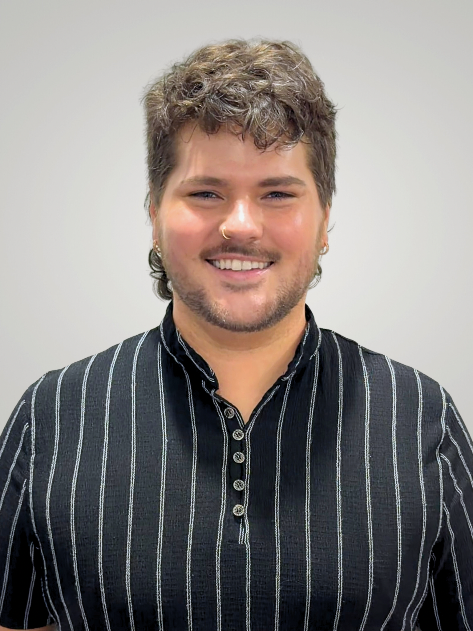 A young person with short, curly brown hair, wearing a black shirt with white vertical stripes and buttons, smiling in front of a plain white wall.
