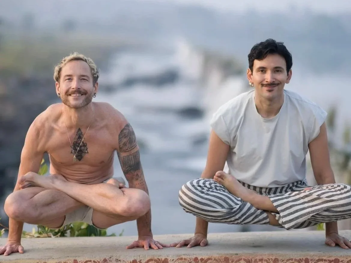 Two men practicing yoga outdoors near water, sitting in a crouched pose with smiles, one shirtless with tattoos and the other wearing a light-colored shirt and striped pants, with boats in the background.