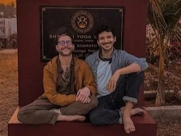Two men sitting cross-legged on a bench in front of a sign, smiling. One is wearing glasses and a brown jacket, the other is wearing a light blue shirt.