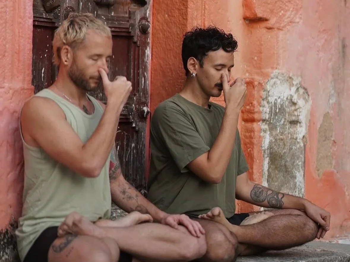 Two young men are sitting cross-legged on the floor with their eyes closed, practicing meditation against an orange wall with peeling paint.