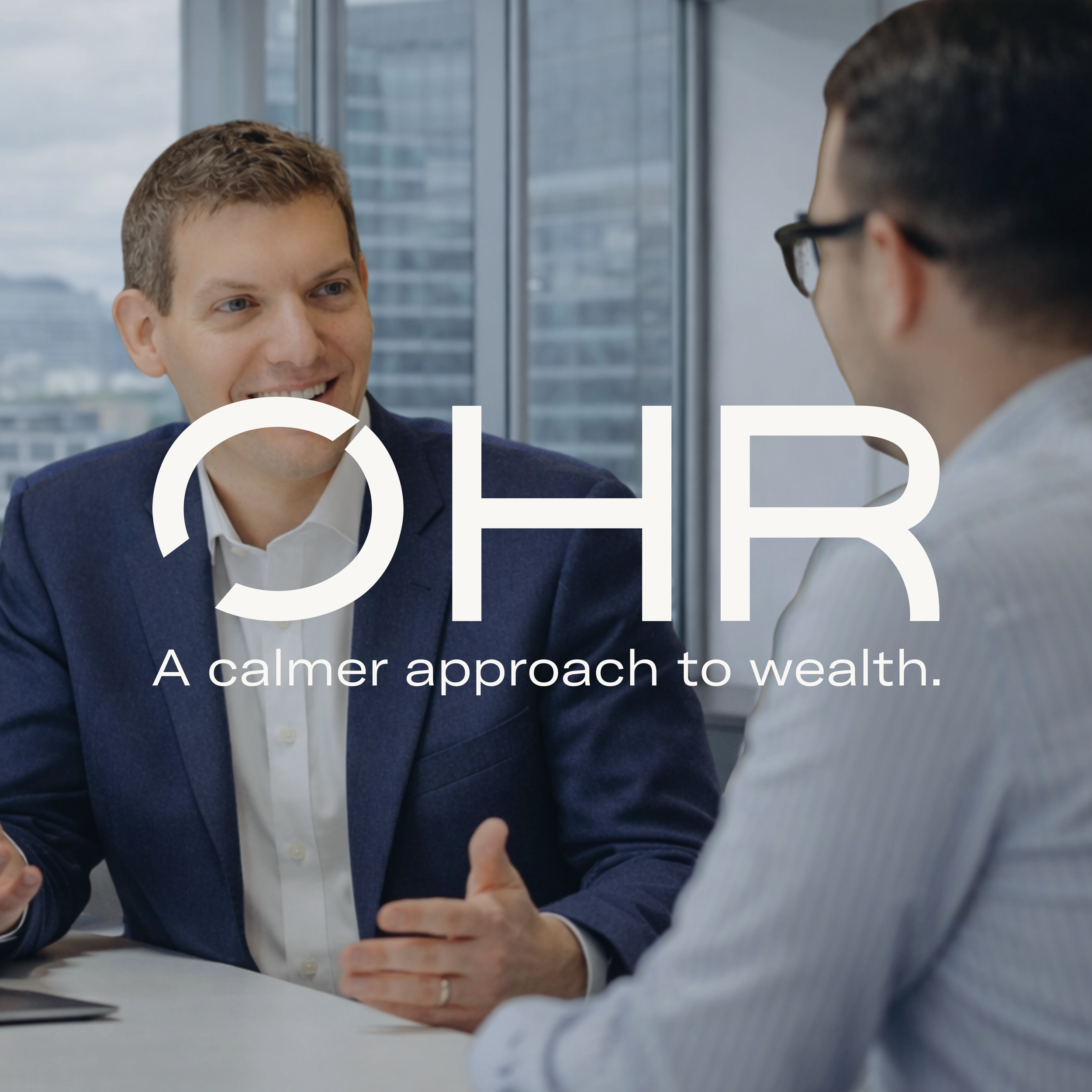 Two men in suits having a conversation in an office with large windows and city buildings in the background.