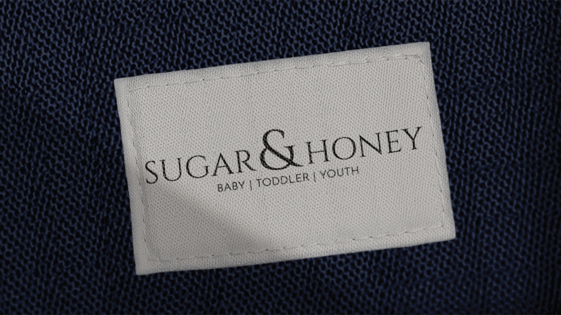Close-up of a fabric label reading 'Sugar & Honey' with the words 'Baby, Toddler, Youth' underneath, sewn onto a textured dark fabric.