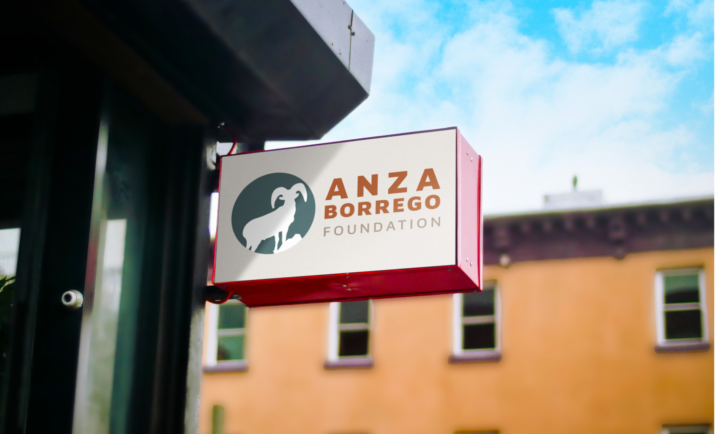 Sign for the ANZA Borrego Foundation mounted on a pole with a cloudy sky and a yellow building in the background.