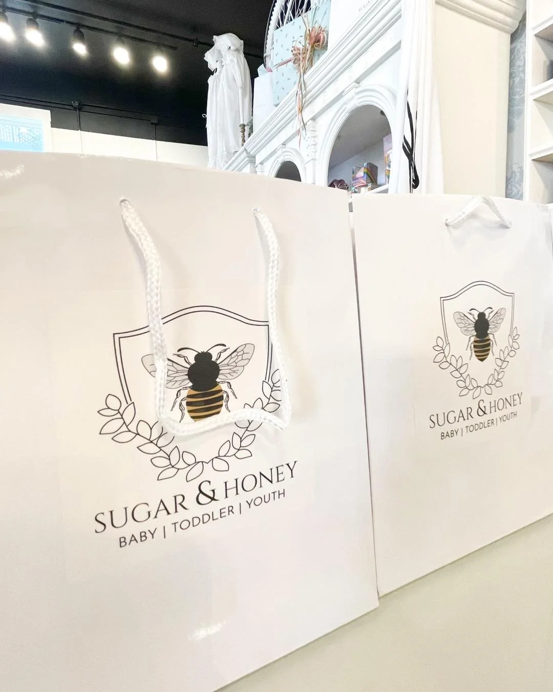 White shopping bags with a bee logo and the words 'Sugar & Honey Baby | Toddler | Youth' printed on them, placed on a surface in a retail store.