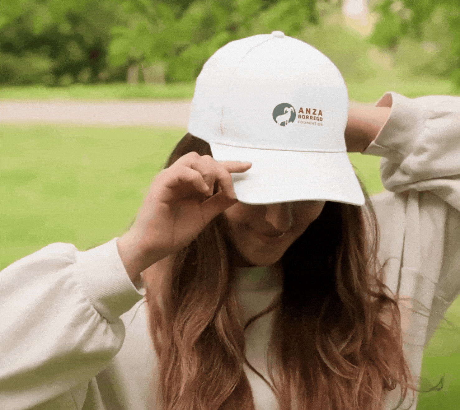 A woman outdoors wearing a white cap with the ANZA Borrego Foundation logo, adjusting the cap while looking down. She has long, wavy reddish-brown hair and is dressed in a light-colored jacket.