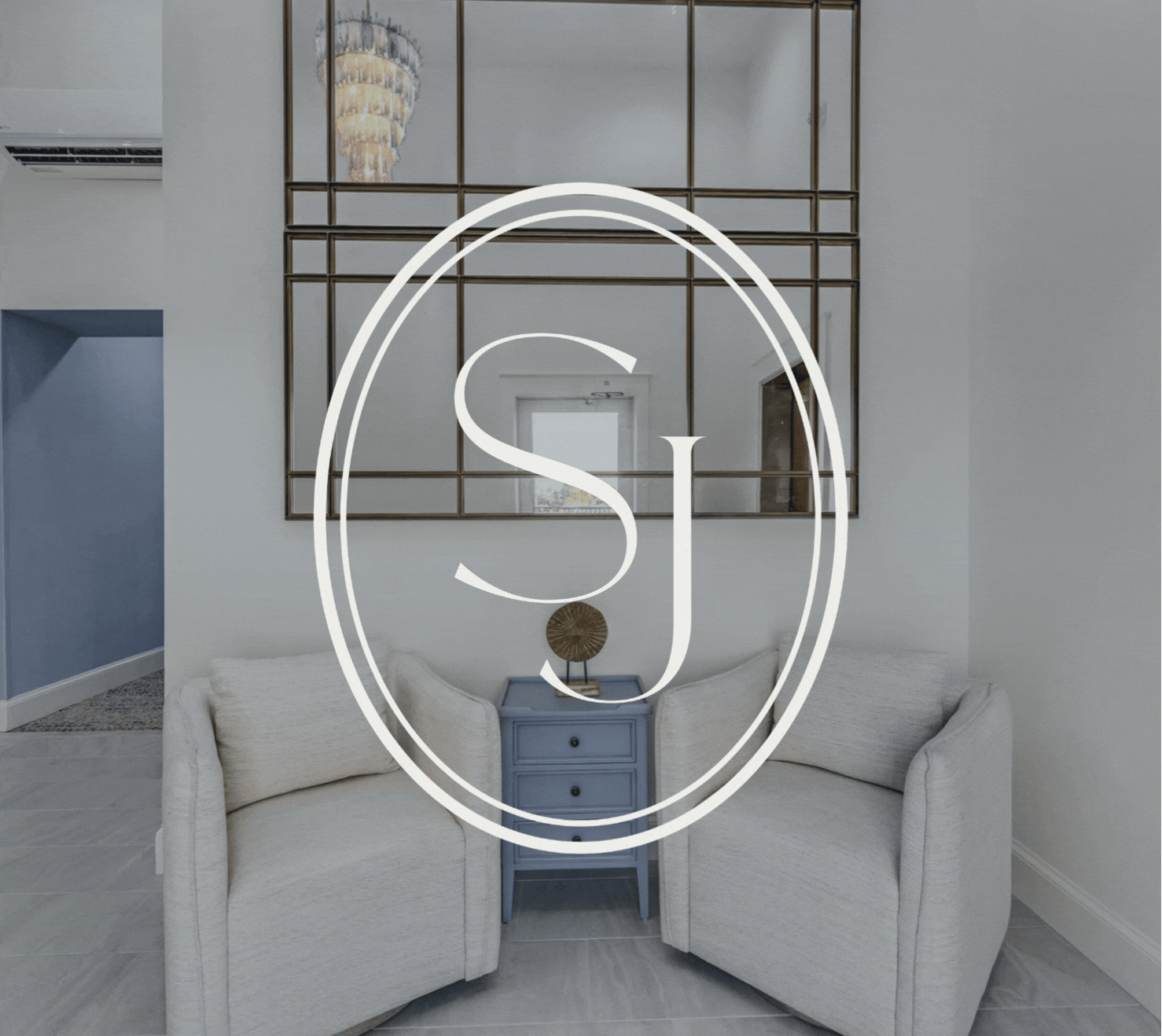 A living room with two white armchairs, a blue side table with drawers, a decorative object on the table, a large mirror with a metallic frame on the wall, and a chandelier hanging from the ceiling.