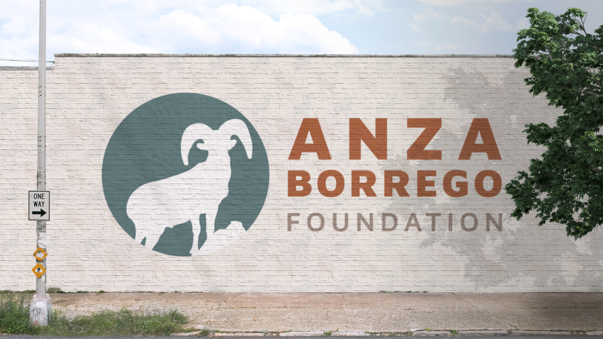 A mural on a brick wall features a circular logo of an animal, possibly a ram, in silhouette within a teal circle. To the right, the text reads 'ANZA BORREGO FOUNDATION' in large brown and grey letters. A one-way street sign with an arrow points to the right is visible on the left side of the wall, and there are trees and a cloudy sky above.