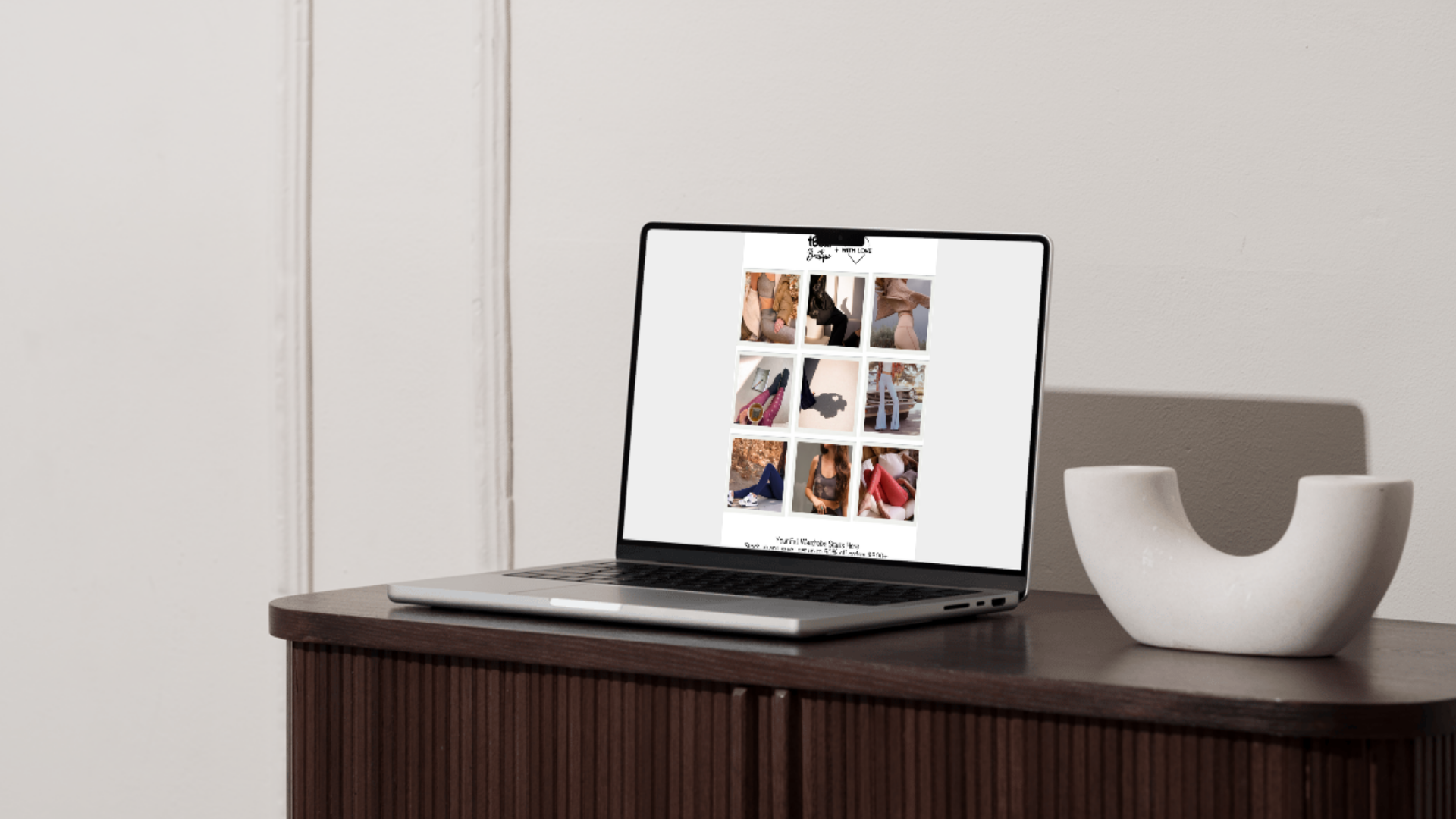 Laptop on a dark wooden table displaying a grid of images, with a white decorative object beside it, against a plain light-colored wall.