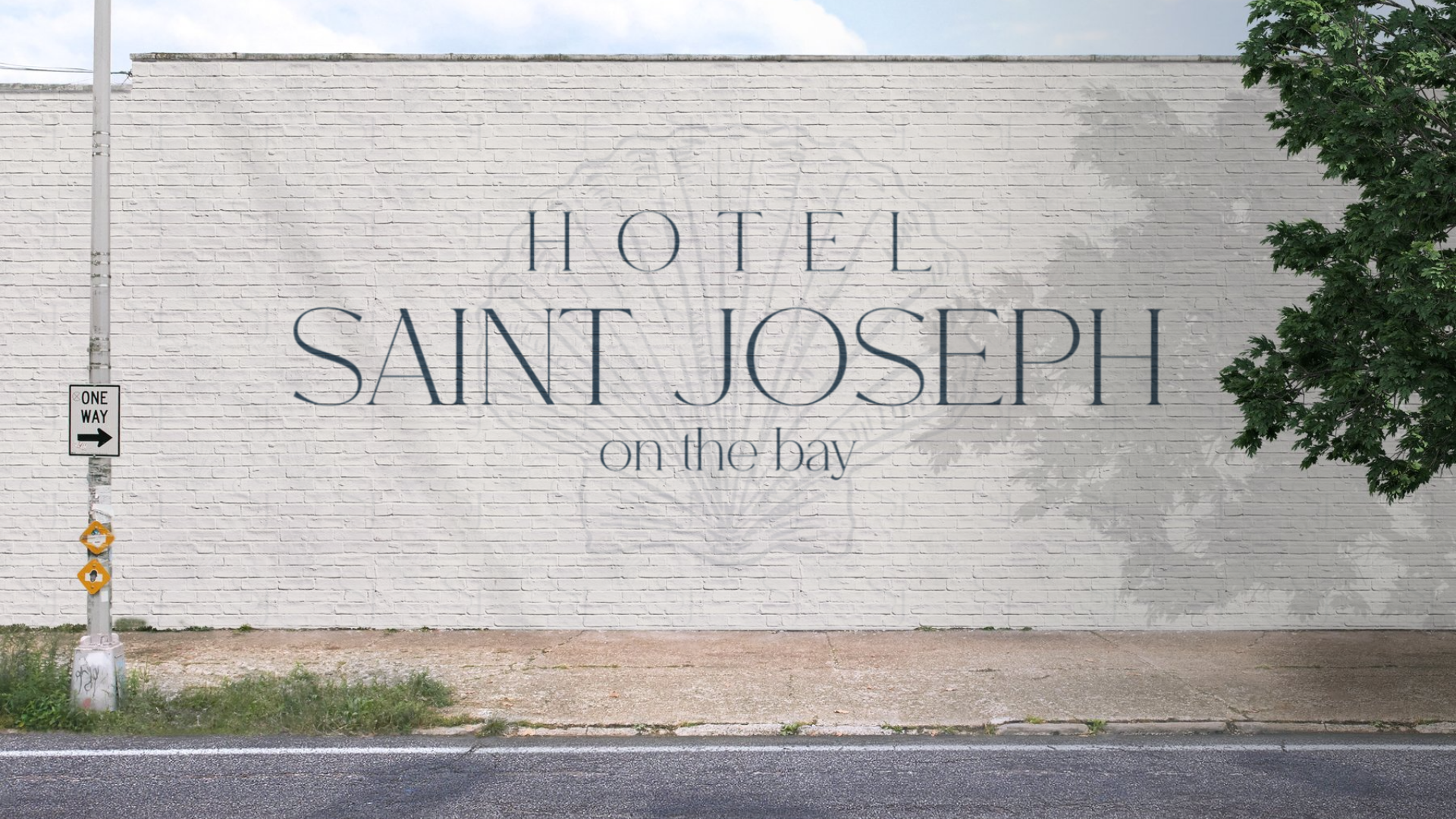 Large white brick wall with painted sign saying 'Hotel Saint Joseph on the bay' and a faint illustration of a compass rose behind the text. A tree is on the right, and a sign with an arrow points right on a pole on the left.