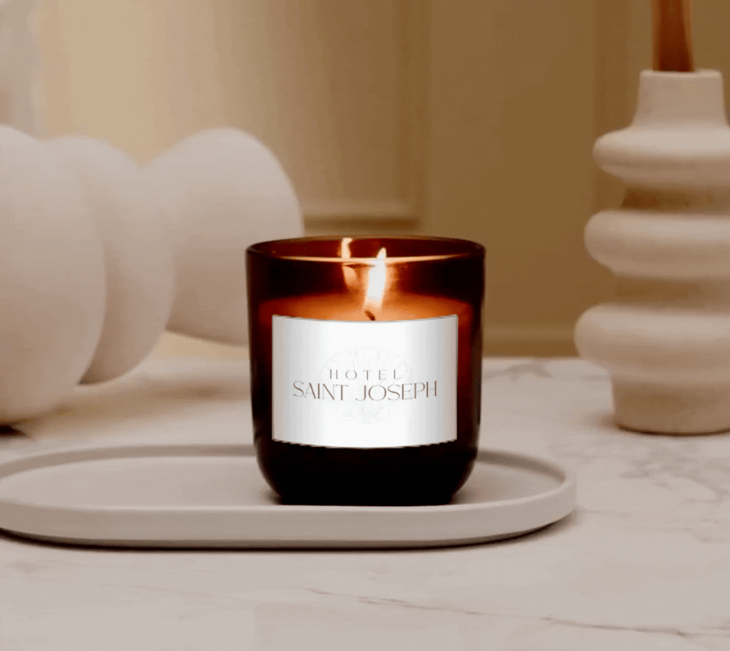 A lit candle in a brown glass holder with a white label reading "Hotel Saint Joseph" is on a white tray, with decorative white objects on either side.