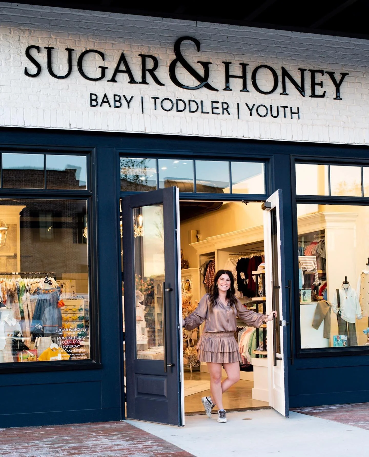 Woman in a brown dress smiling and standing in the doorway of a children's clothing store called Sugar & Honey.