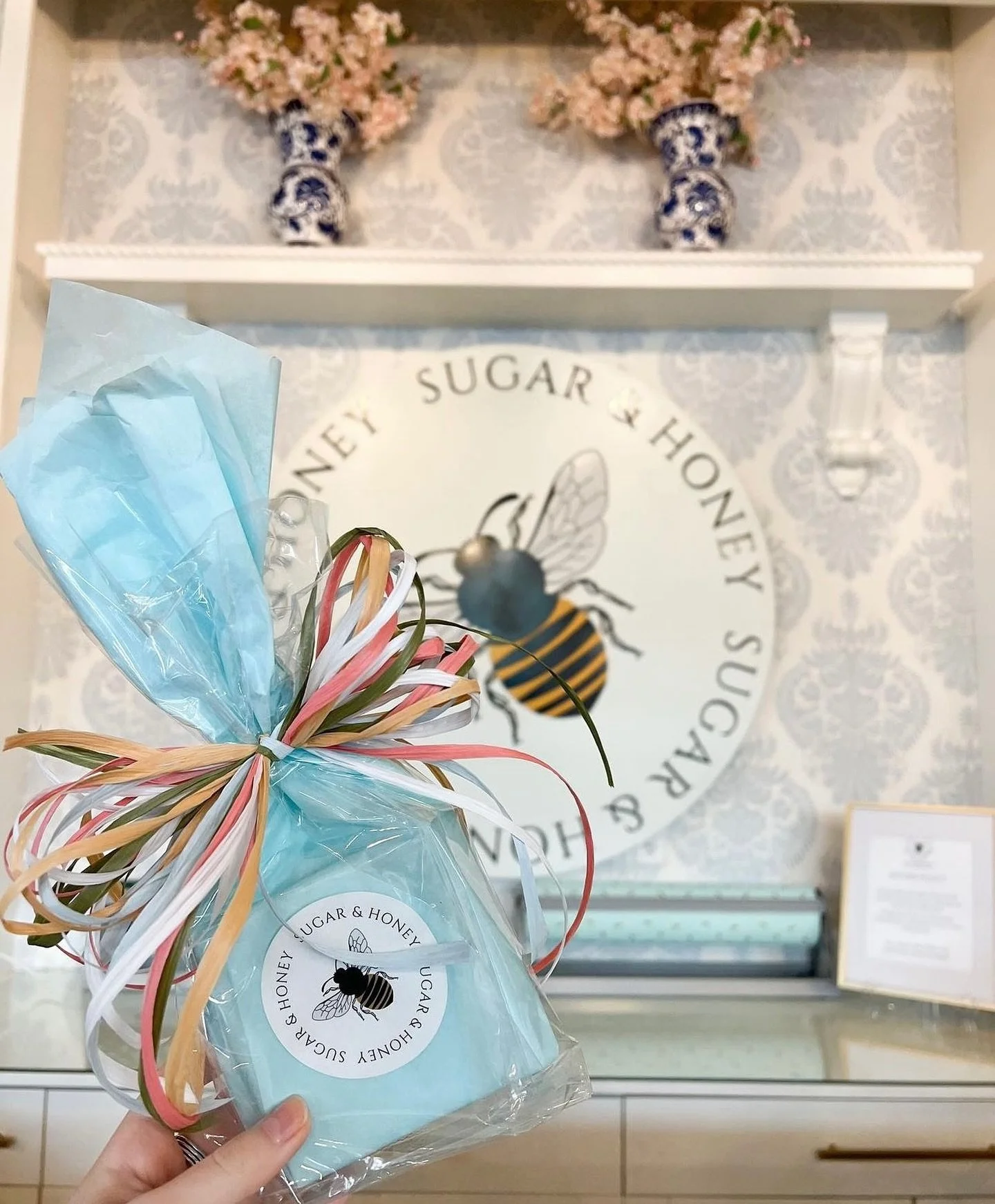 Person holding a gift bag wrapped in light blue tissue paper and colorful ribbons, in front of a wall with honey bee logo and 'SUGAR & HONEY' text, with decorative vases and flowers on a shelf in the background.