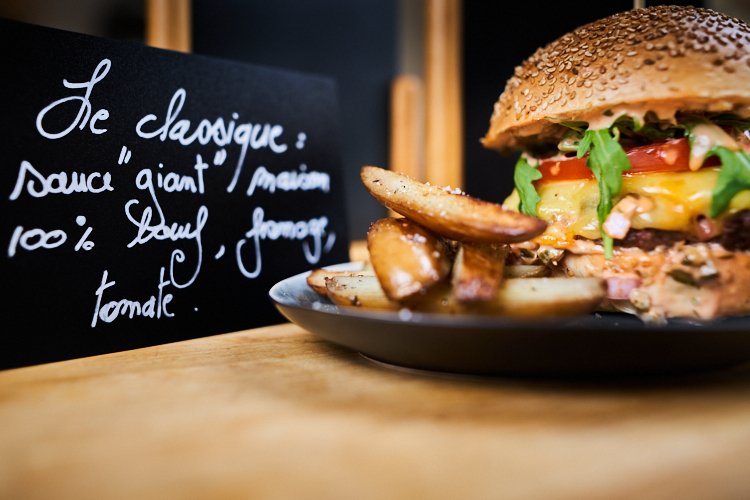 Close-up of a cheeseburger with lettuce, tomato, and sauce, served with potato wedges on a black plate. A chalkboard with French writing is in the background.