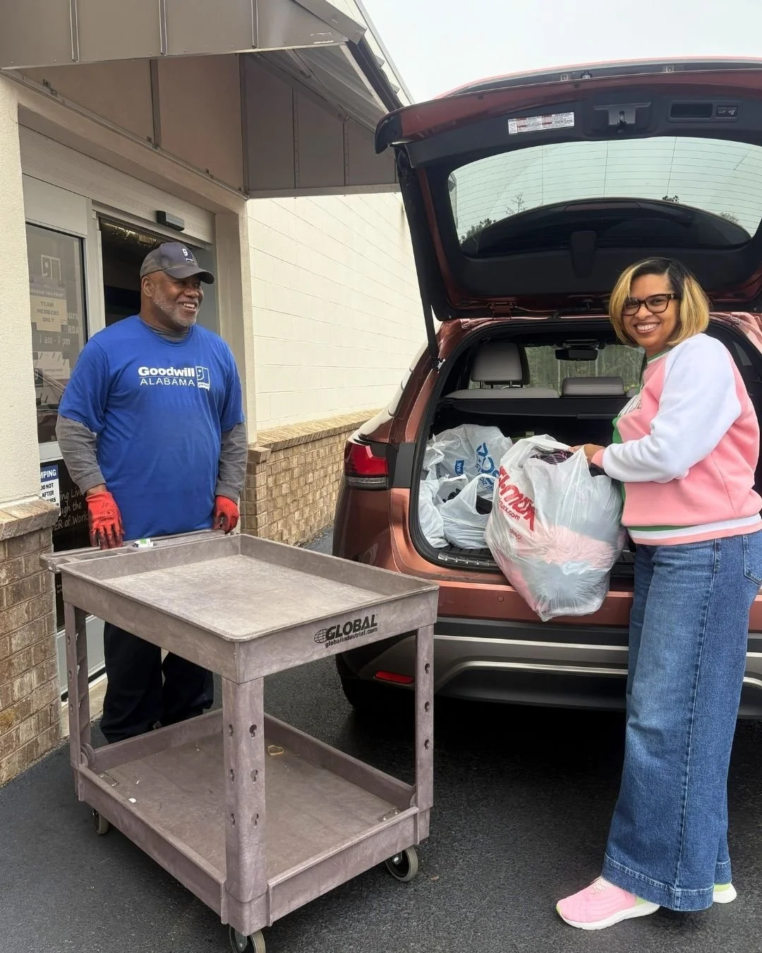 A woman is loading groceries into the back of a parked car while a man stands nearby, smiling. The man is wearing a blue Goodwill Alabama t-shirt and gloves, and there is a metal cart in front of him.