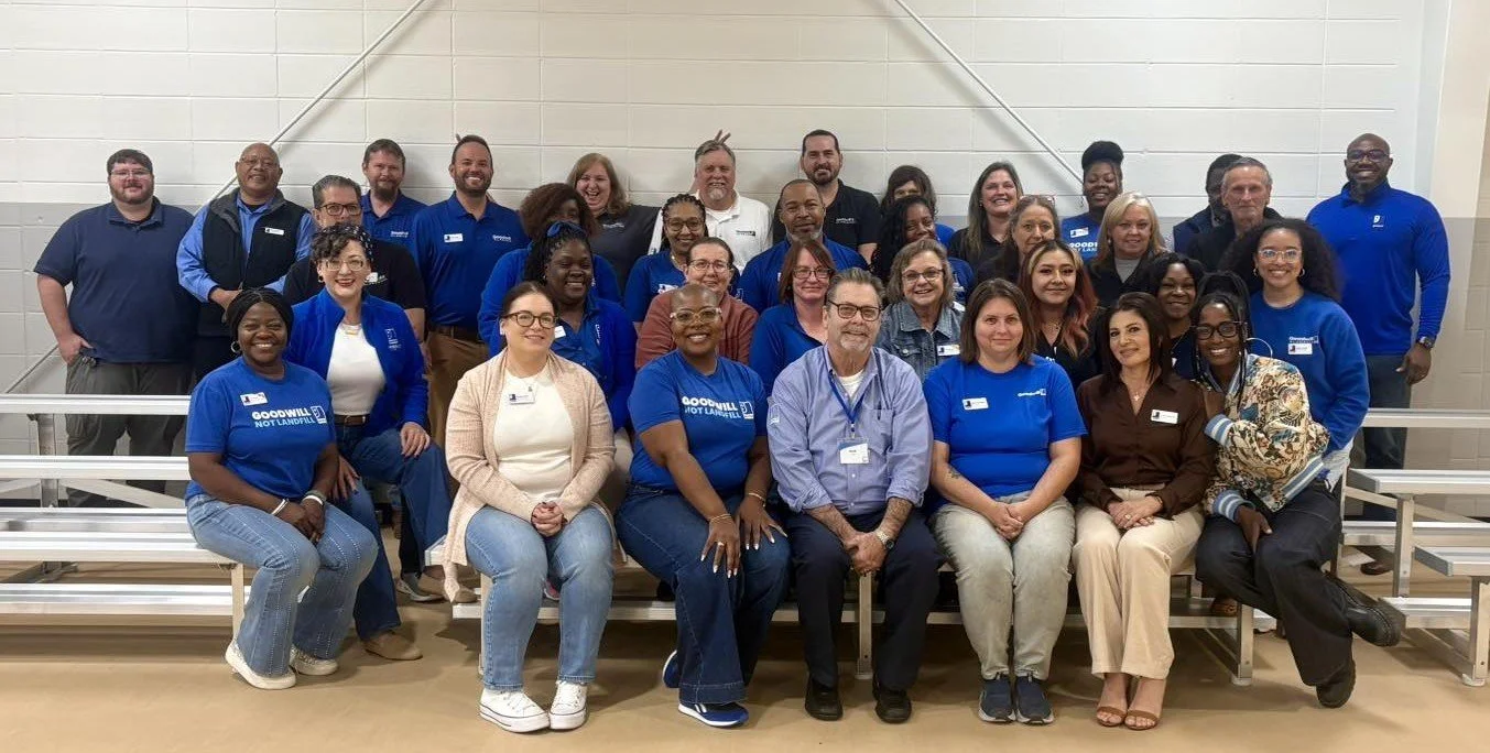 Group photo of diverse people, mostly women, gathered in a gymnasium with bleachers, some wearing blue shirts and holding microphones, smiling for the camera.