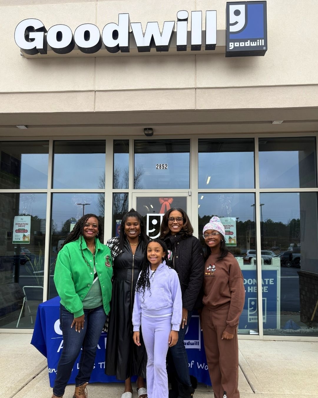 Five women and a girl standing outside a Goodwill store, smiling, with donation drive signs visible behind them.