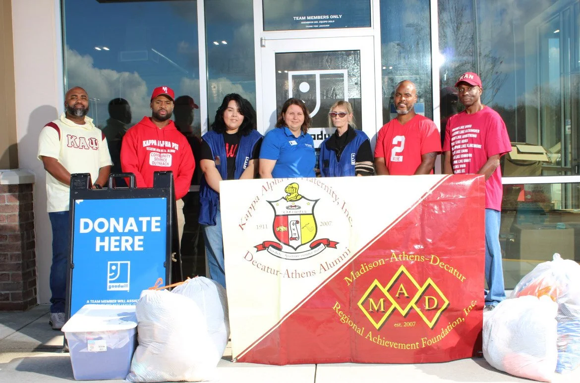 Group of people standing outside a building with donation bags, a sign that says 'Donate Here,' and banners for Alpha Phi Alpha and Madison-Athens Decatur Regional Achievement Foundation, Inc.