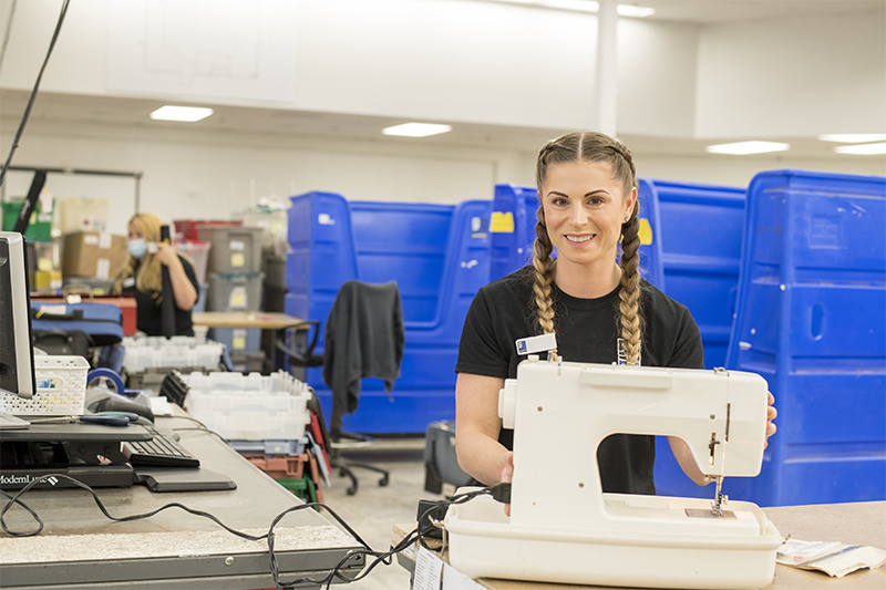 A woman smiling and wearing a blue vest and cap in a clothing sorting warehouse, with other workers in white shirts and masks, and shelves filled with shoes and other items.