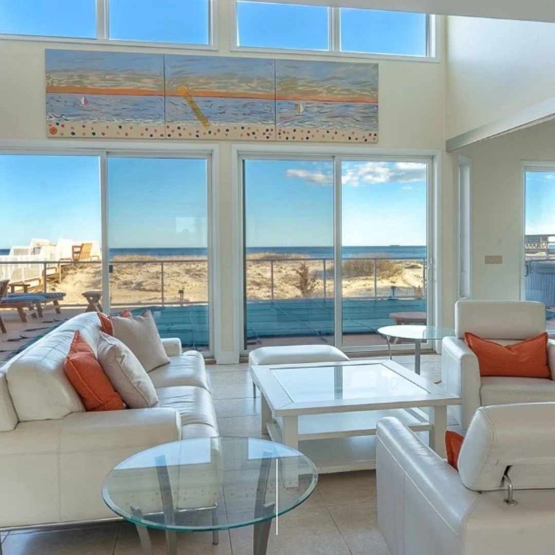 Living room with white couches and orange pillows, large glass sliding doors revealing a beach and ocean view, and a painting of a beach scene above the doors.