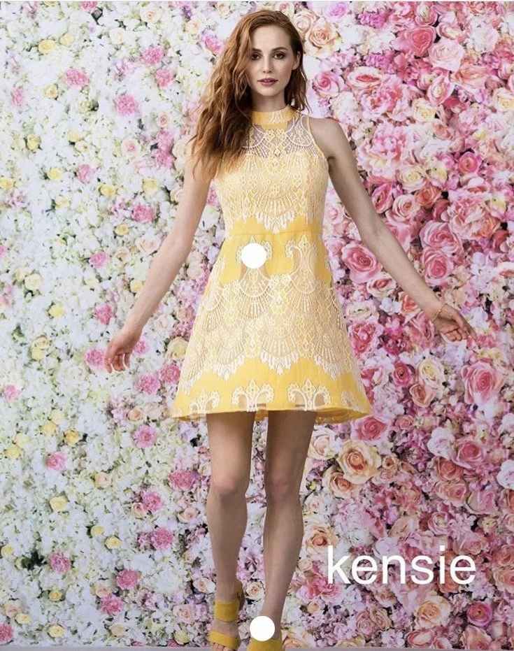 A woman wearing a yellow lace dress standing in front of a floral pink and white background.