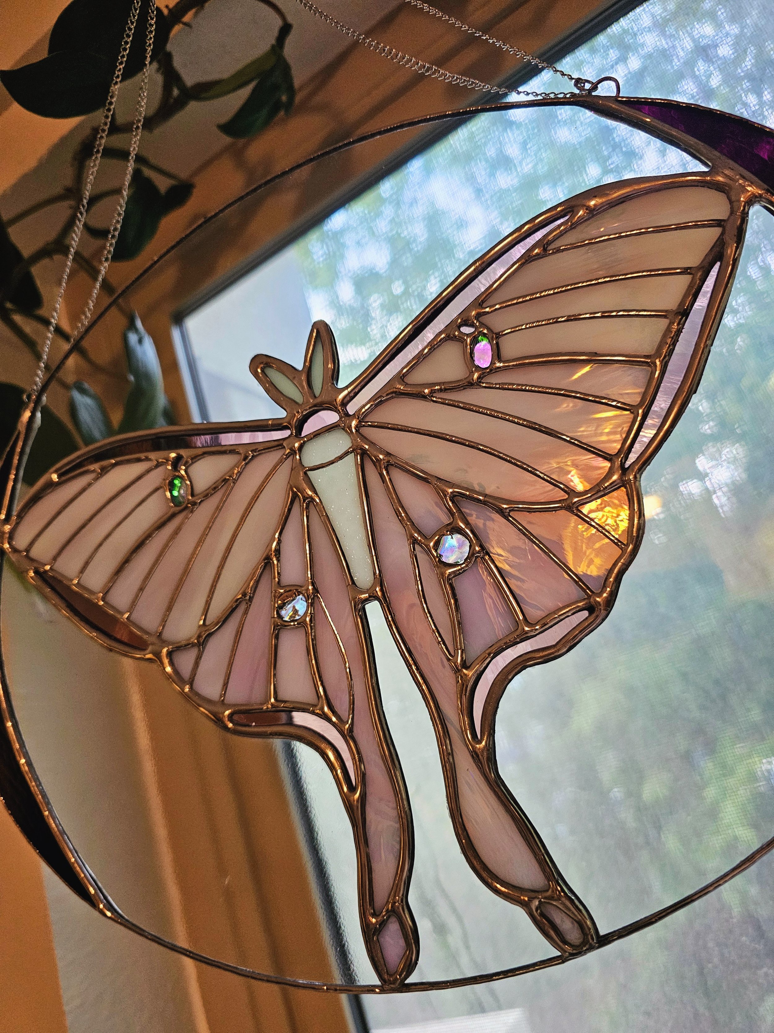 A butterfly-shaped stained glass ornament with pink, purple, green, and iridescent glass pieces, hanging from a chain near a window, with a blurred view of greenery outside.