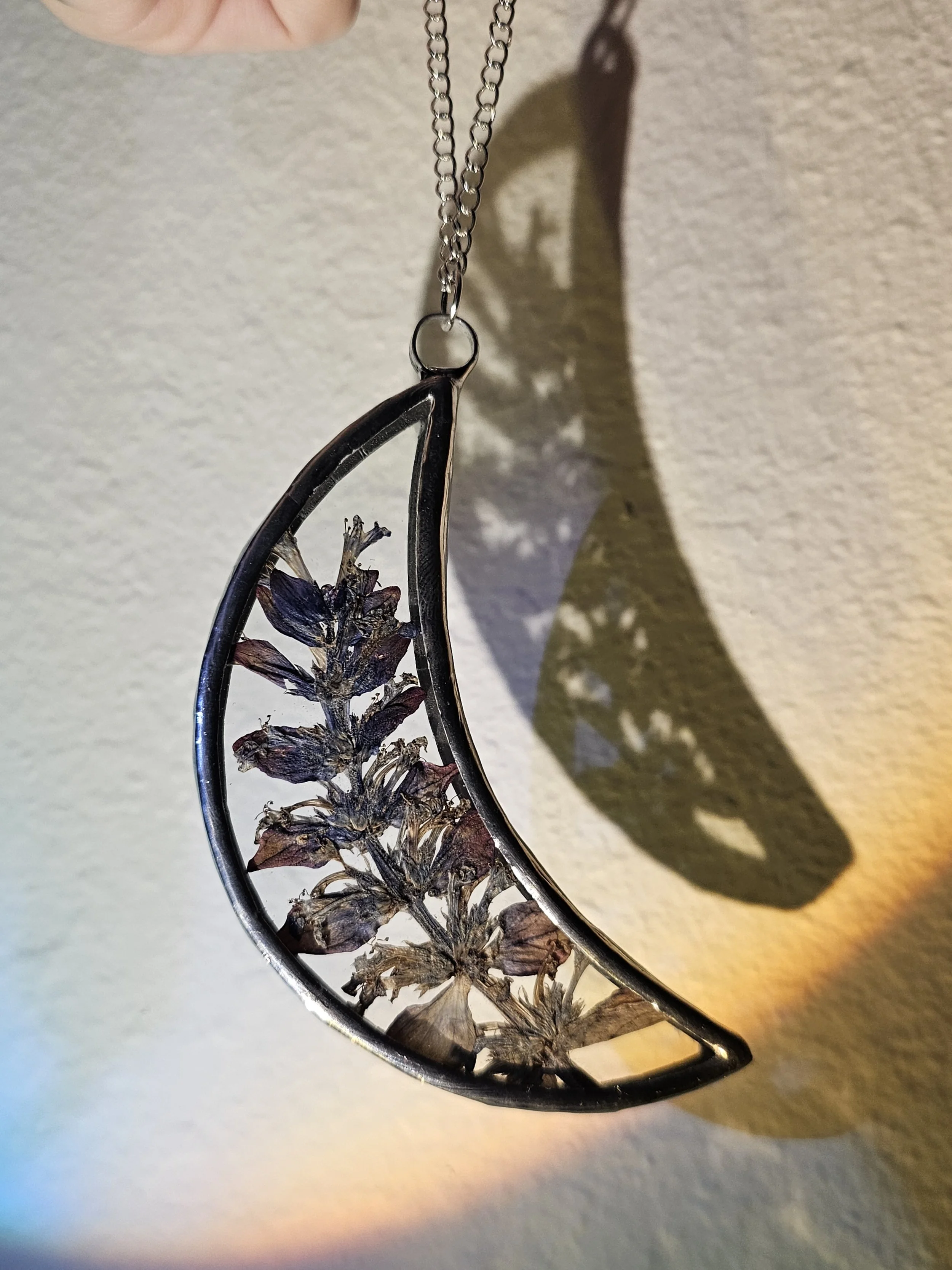 Close-up of a metal necklace with a dried flower inside its open teardrop-shaped frame, hanging against a neutral background with a shadow cast by the necklace.