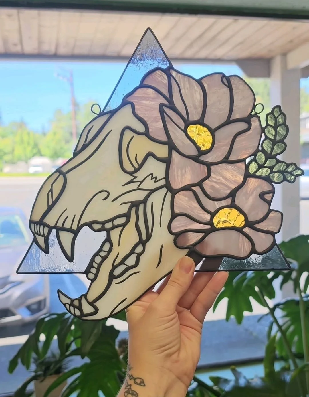 Hand holding a stained glass artwork of a dinosaur skull with flowers and leaves, held up against a window with trees and sky outside.