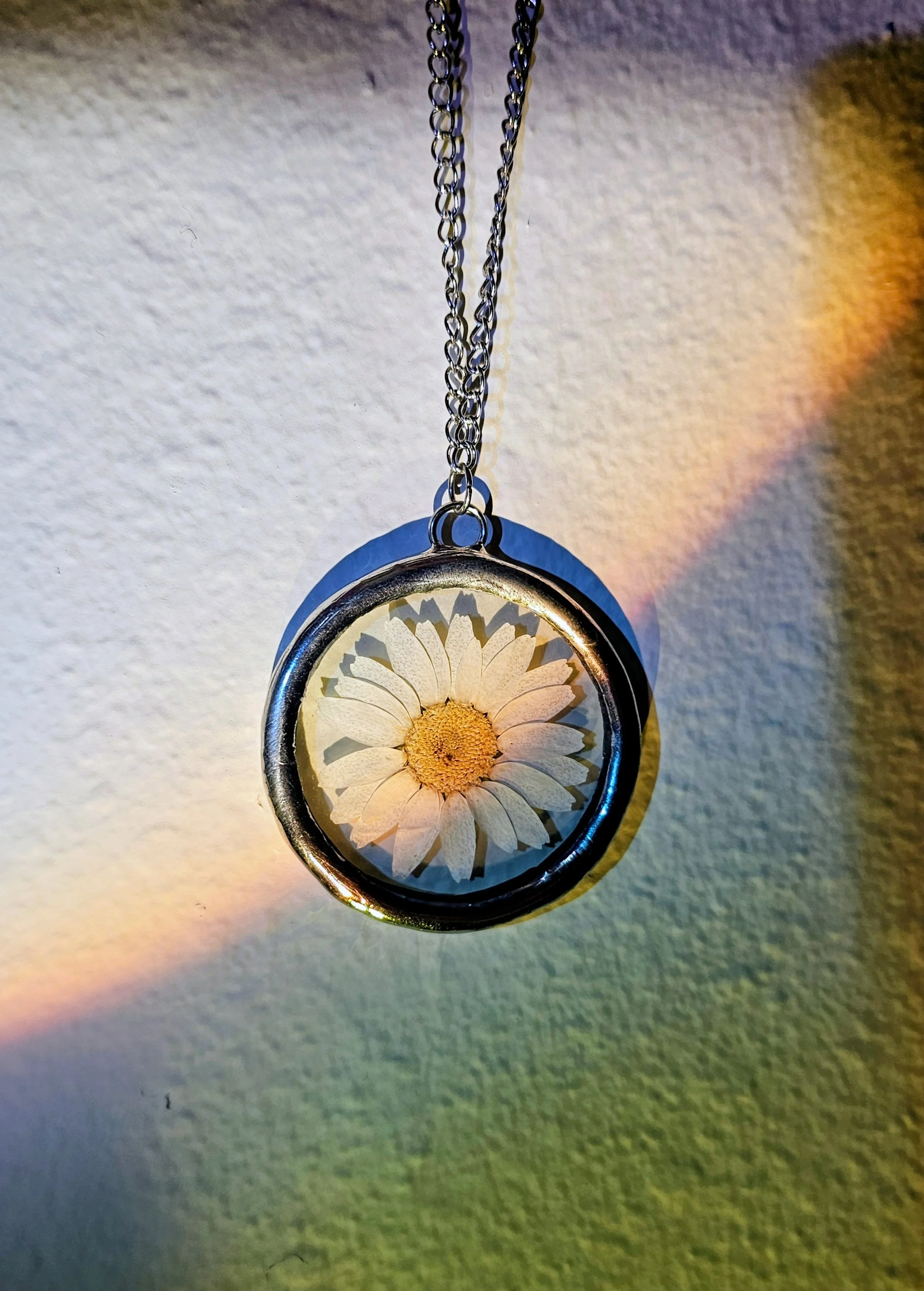 A silver pendant necklace hanging from a chain against a textured white wall, with a daisy flower inside the pendant.
