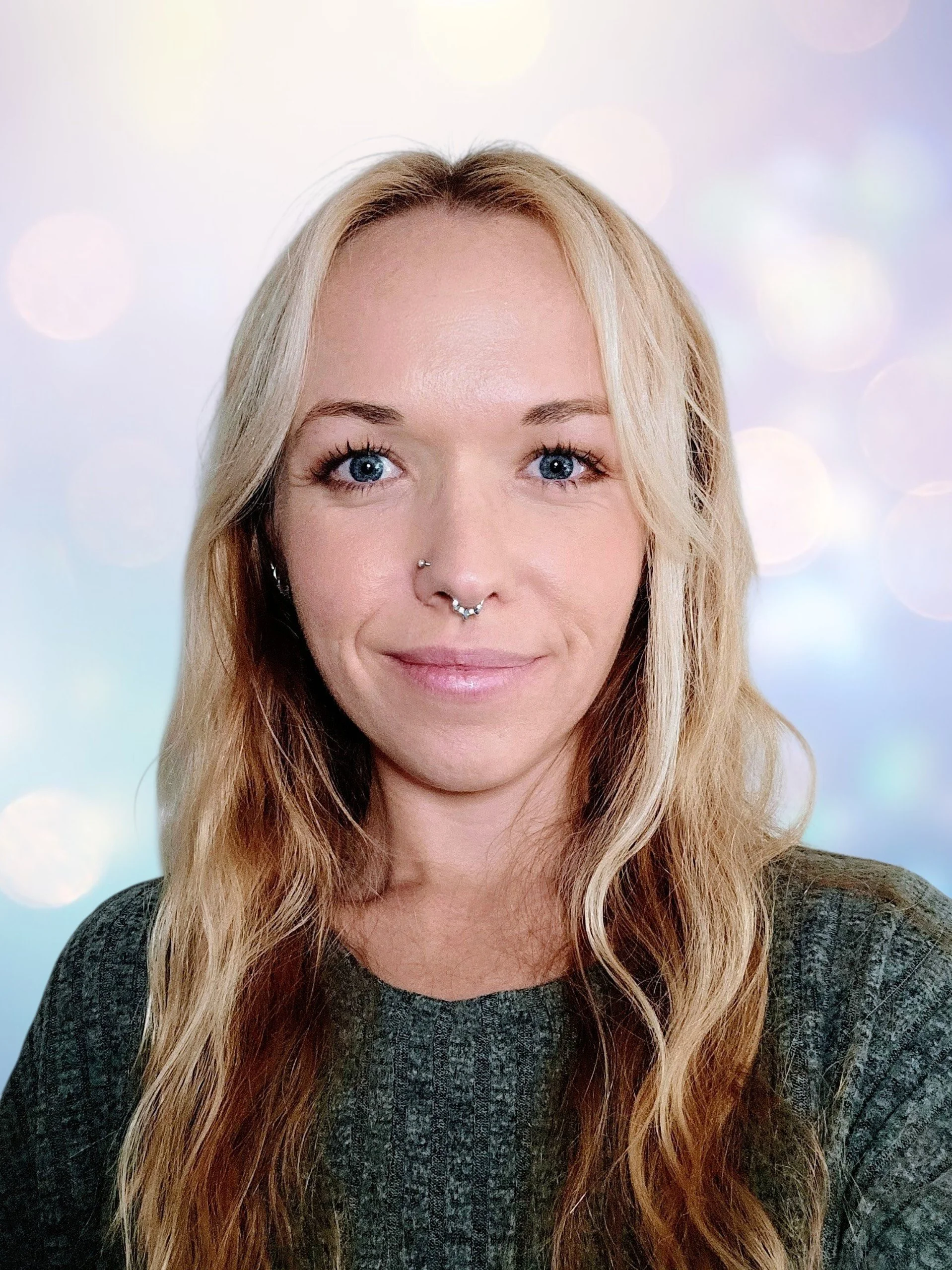 Close-up of a woman with long blonde hair, blue eyes, and a septum piercing, wearing a dark sweater, smiling softly against a blurred, pastel-colored background.
