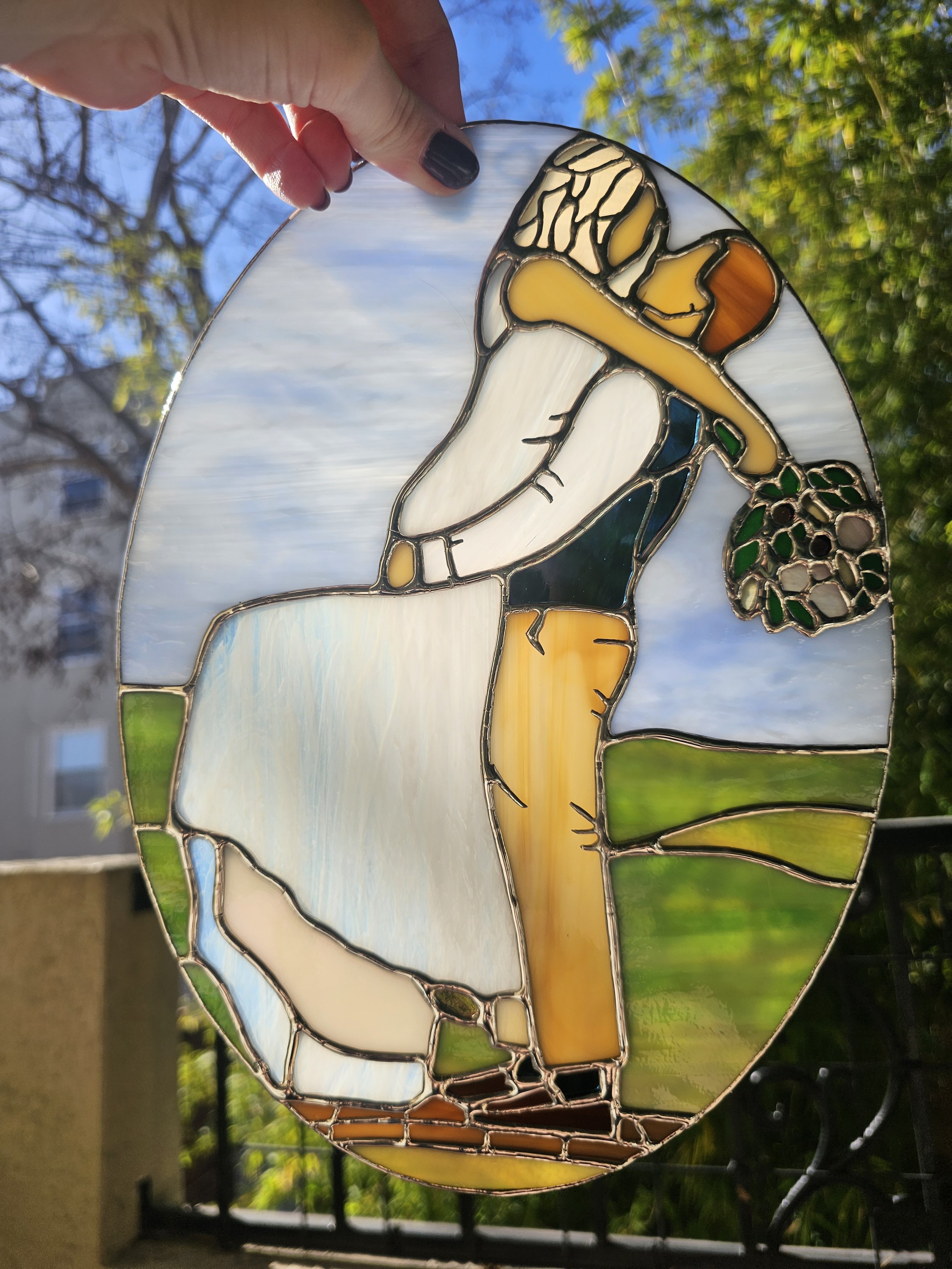 A stained glass art piece in the shape of a dog, with colorful sections of glass depicting different parts of the dog, held up against a sunny outdoor background with trees and a building.