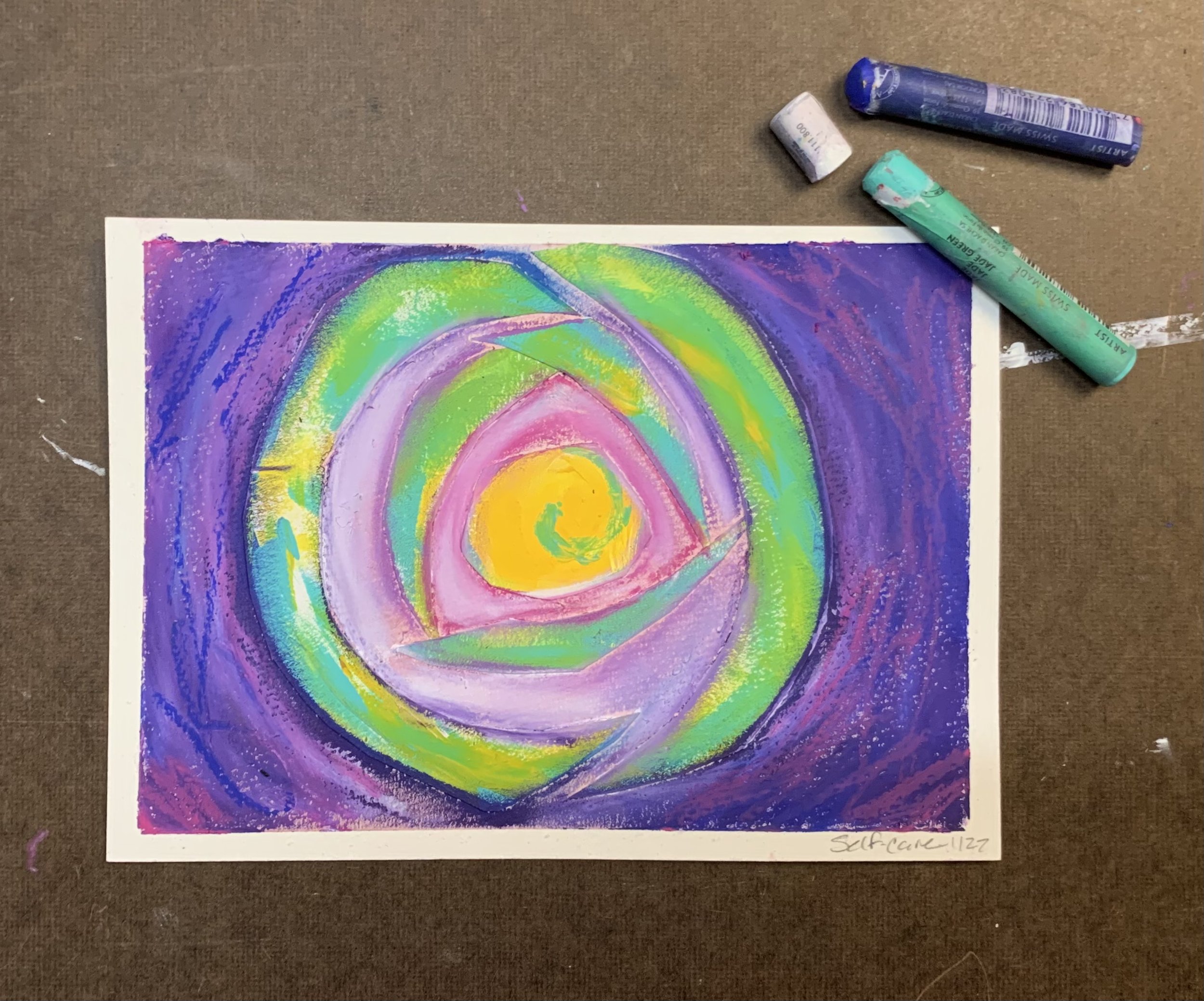 Abstract pastel artwork with colorful spiral design. Bright hues of yellow, green, pink, and purple blend together. Pastel sticks are beside the artwork.