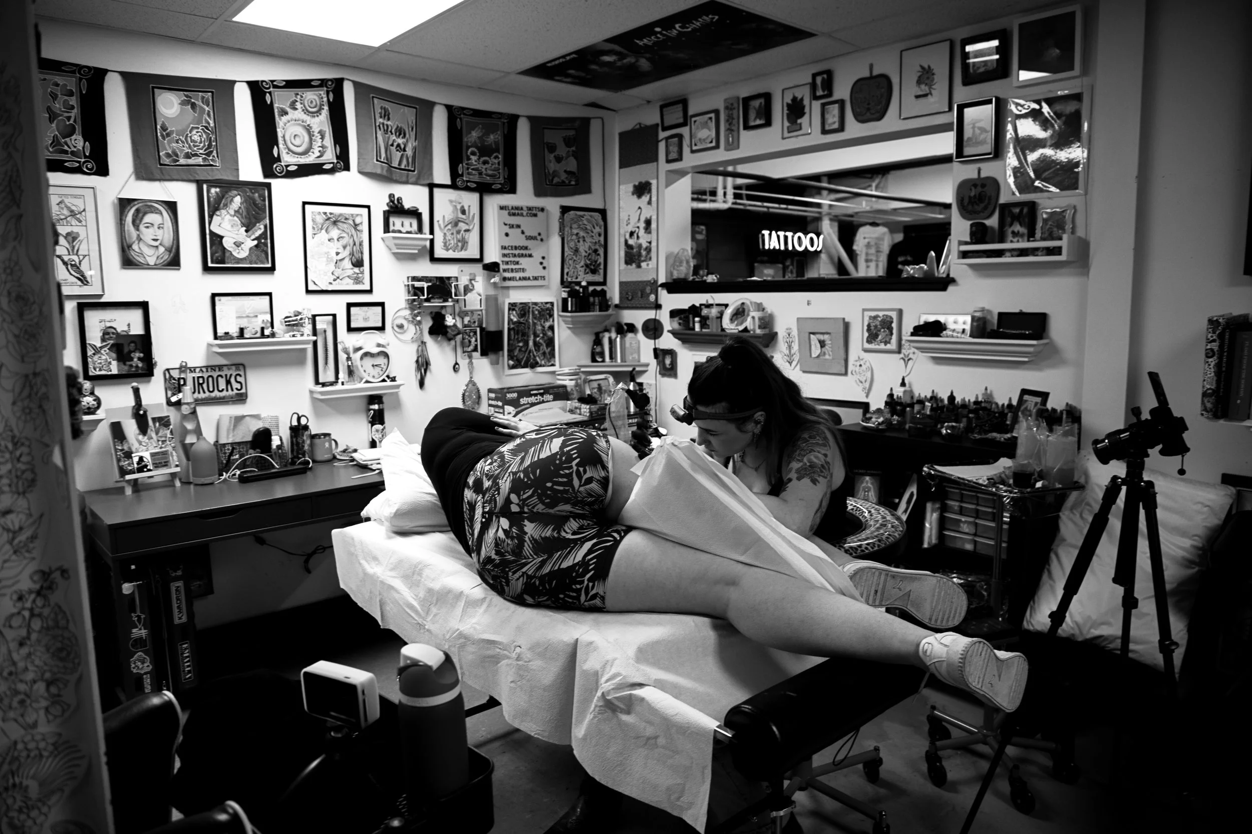 A woman is lying on a table getting a tattoo, with her leg elevated. The room is decorated with framed artwork, posters, and miscellaneous items on shelves. A tattoo artist is working on her leg, and various tattoo supplies are visible on nearby surf