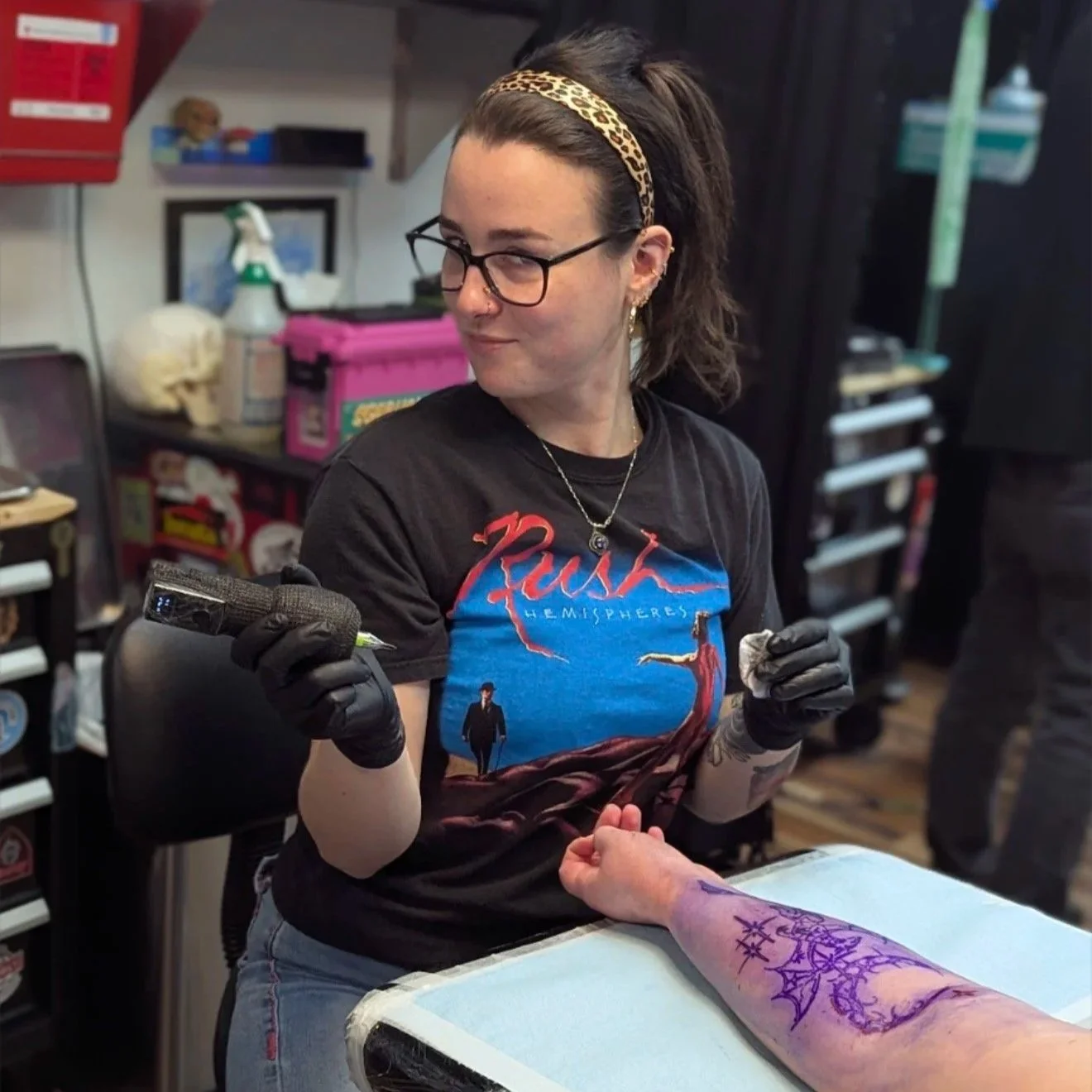A woman with glasses and a leopard-print headband tattooing a person's arm in a tattoo shop. She is wearing a Rush band t-shirt and gloves, holding tattoo equipment, with tattoo design on the person's arm.