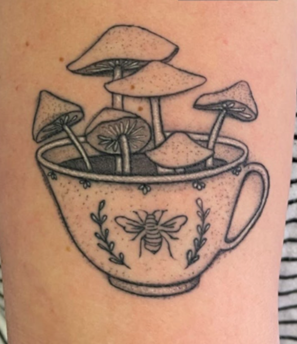 A tattoo of a teacup with mushrooms growing inside and a bee symbol on the front, with the word 'Healed' above it.