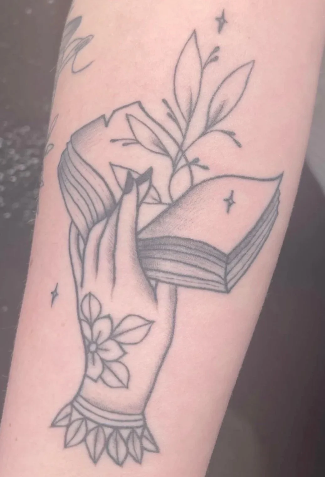 Tattoo of a hand holding an open book with leaves and stars around.
