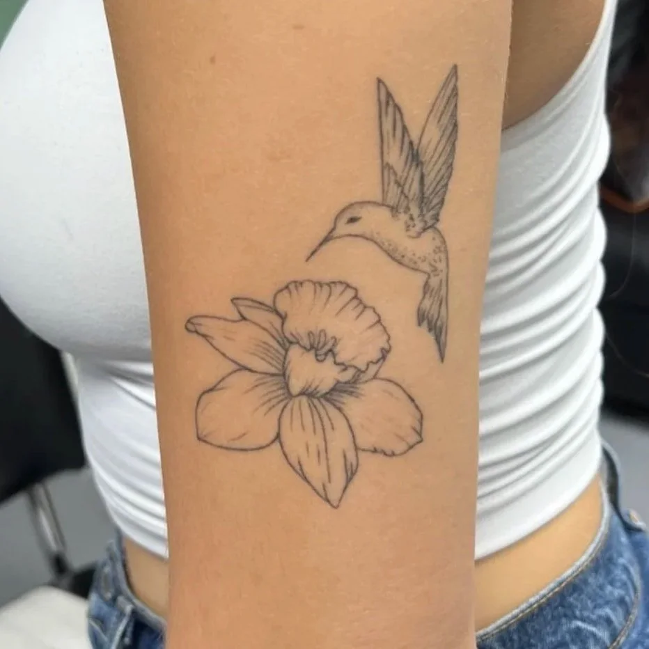 A woman with a tattoo of a hummingbird and a flower on her upper arm. The tattoo is black and gray line art. The word "Healed" is overlaid on the image.