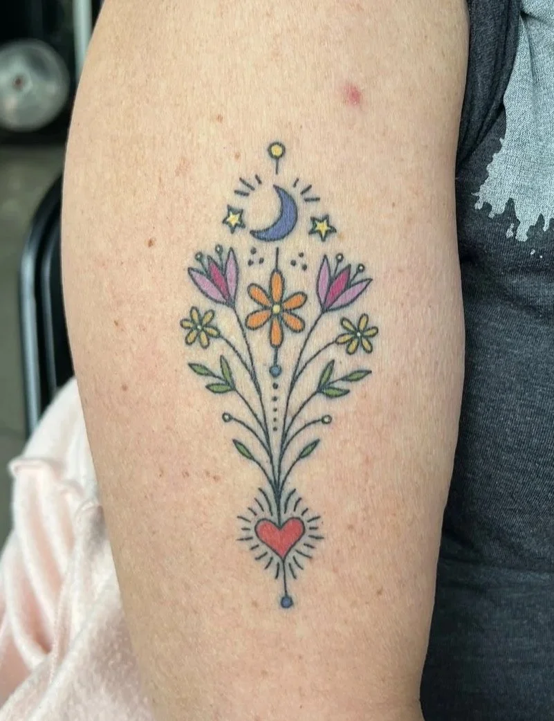 Colorful tattoo of flowers with a moon, stars, and hearts on a person's upper arm.