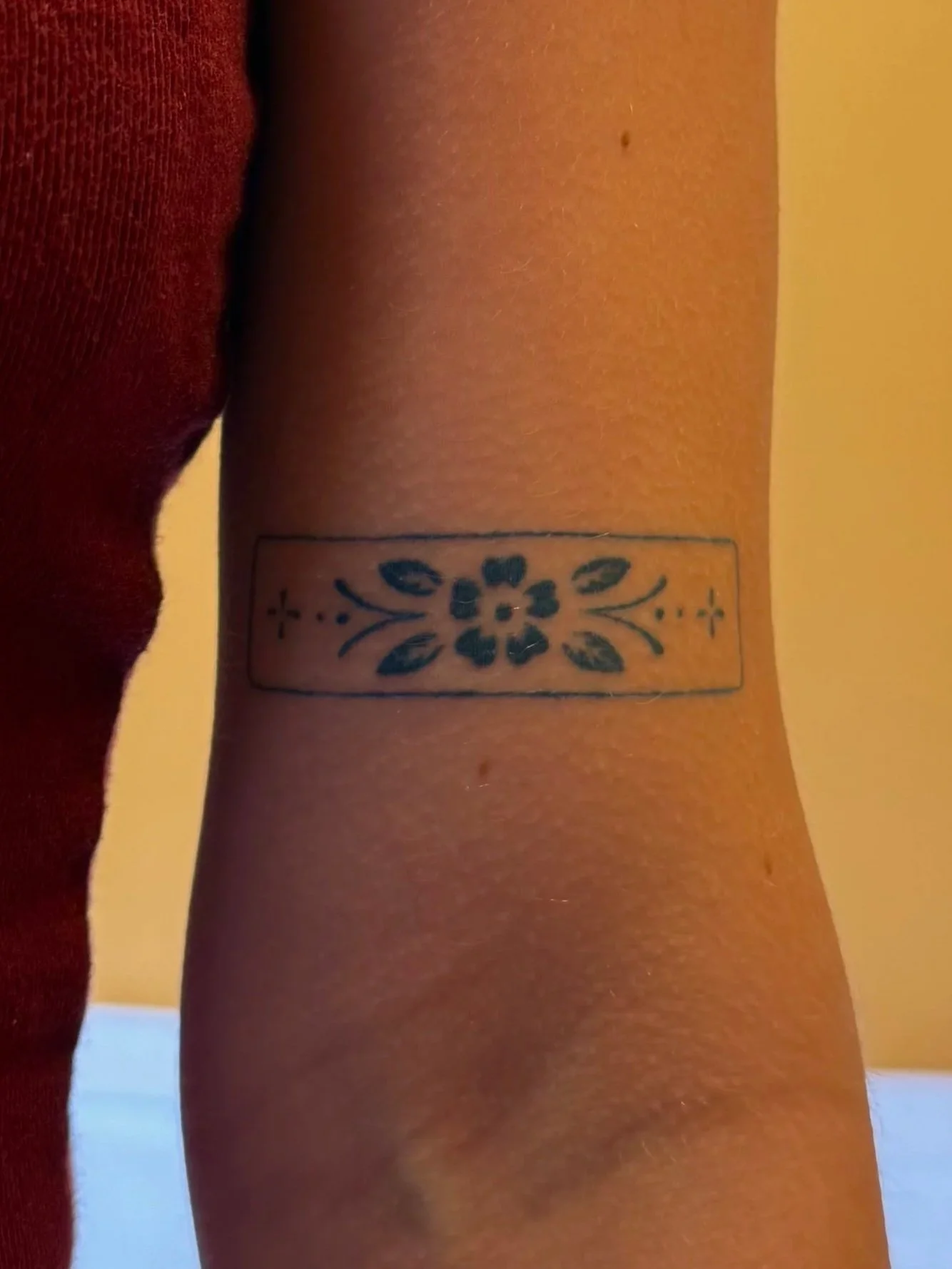Close-up of a person's arm with a rectangular tattoo of a flower design on the inner forearm, wearing a maroon shirt.