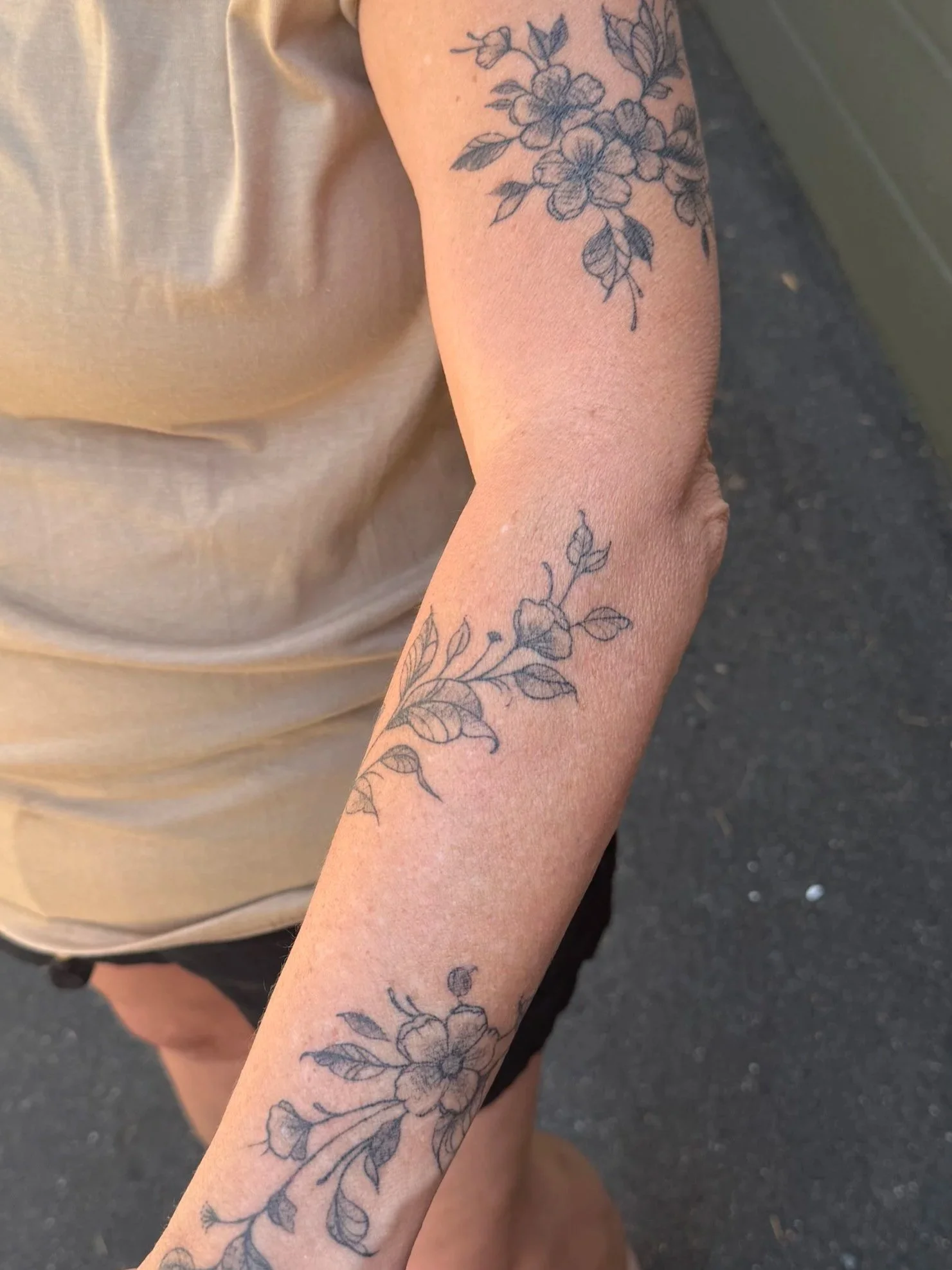 A woman with a tattoo on her arm showing a floral design, standing outdoors near a building with green siding, wearing a beige shirt and a silver necklace with a moon pendant.