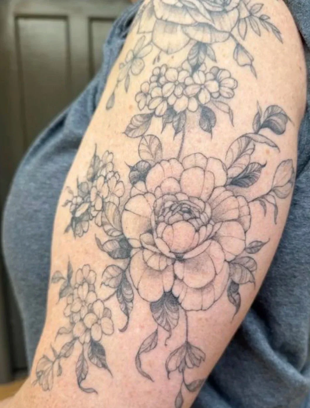 Close-up of a floral tattoo featuring a large flower, possibly a peony, surrounded by smaller flowers and leaves on a person's upper arm.