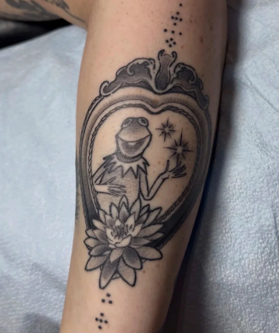 Tattoo of Kermit the Frog with stars and a lotus flower on a person's arm.