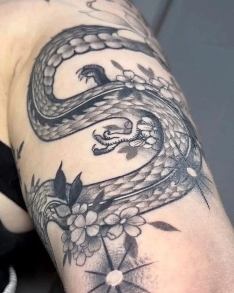 Black and gray tattoo of a dragon surrounded by flowers on a person's upper arm.