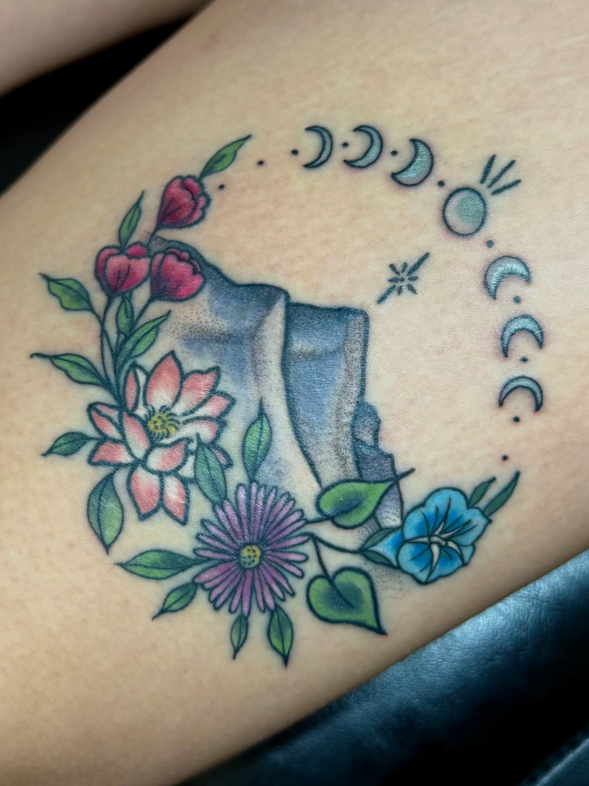 Tattoo of colorful flowers, leaves, and moon phase symbols on skin.