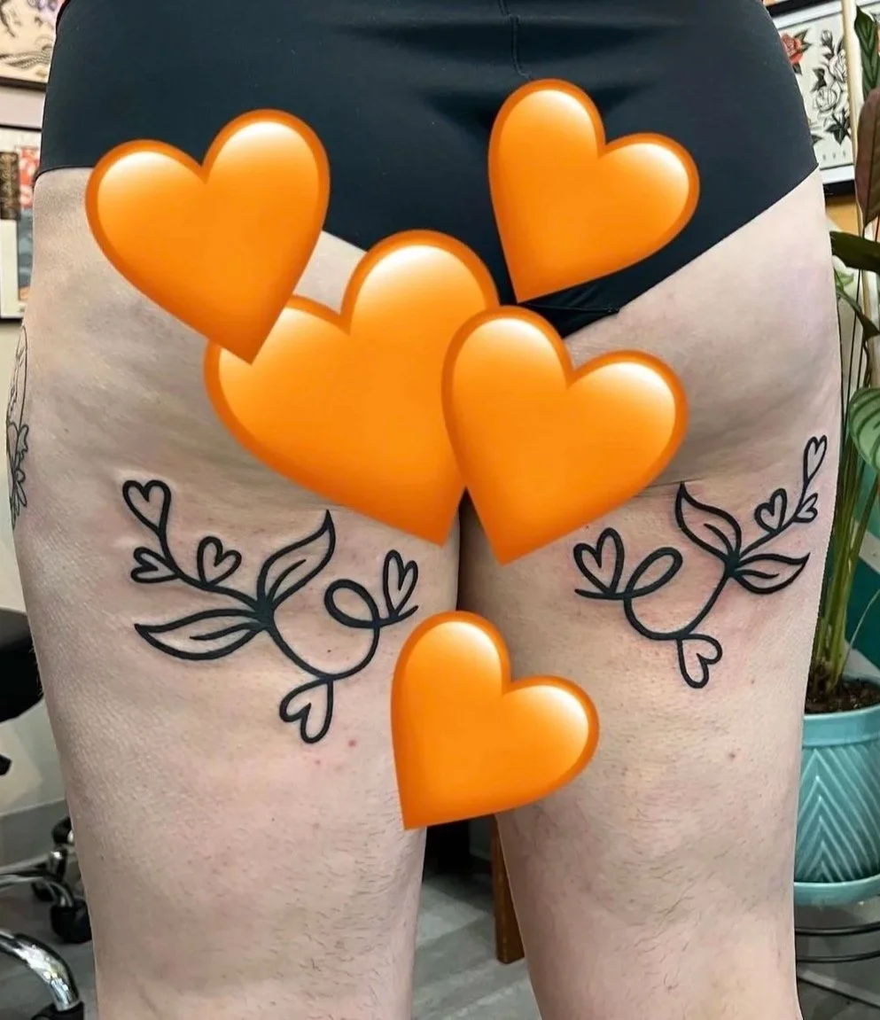 A person wearing black shorts with tattooed arms and decorative hearts and leaves by their thighs, with orange heart emojis over their back and upper thighs.