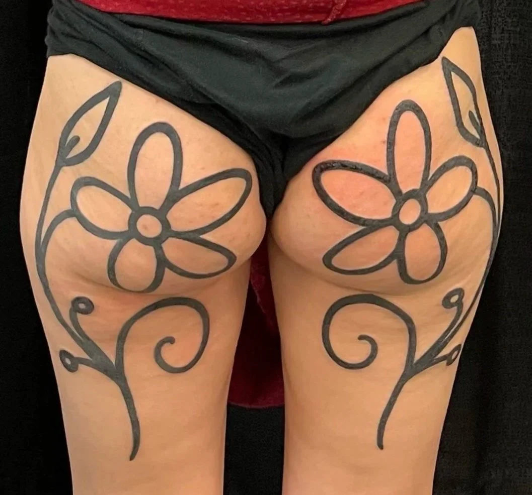 Tattooed floral designs on upper thighs, featuring large flowers with multiple petals and swirling stems in black ink.