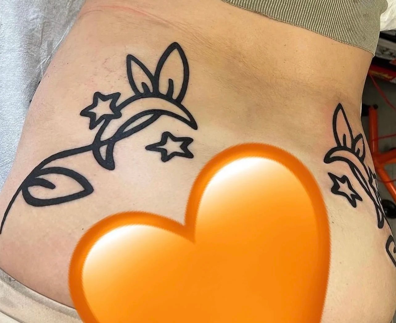 Tattoo of a cartoon-style unicorn face with stars and a crescent moon, located on the person's side near their waist, covered partly by clothing, with an orange heart emoji covering part of the image.