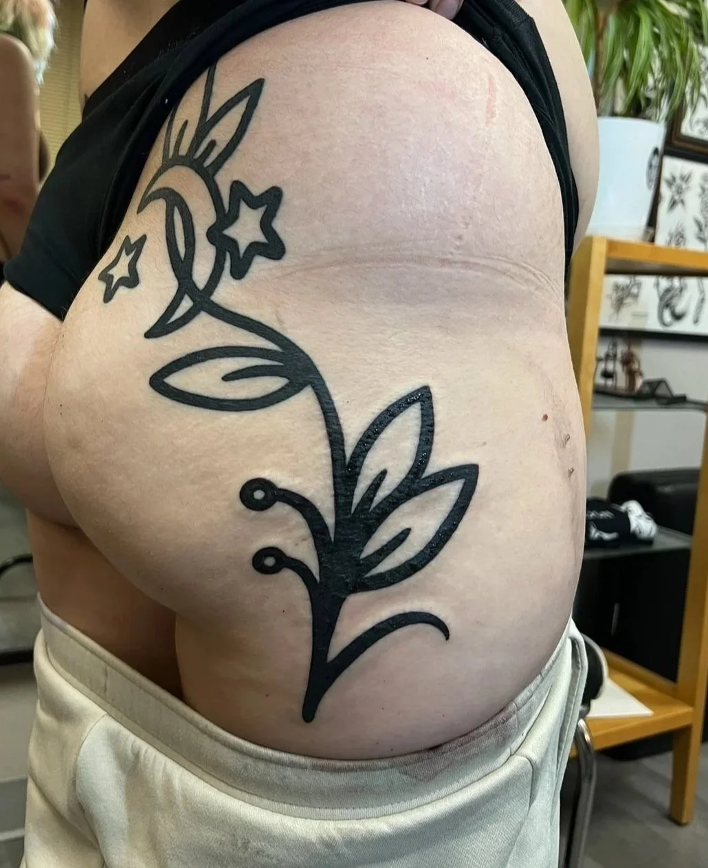 A person's upper arm with a large black outline tattoo of a stylized flower with leaves, star shapes, and a moon with a face.