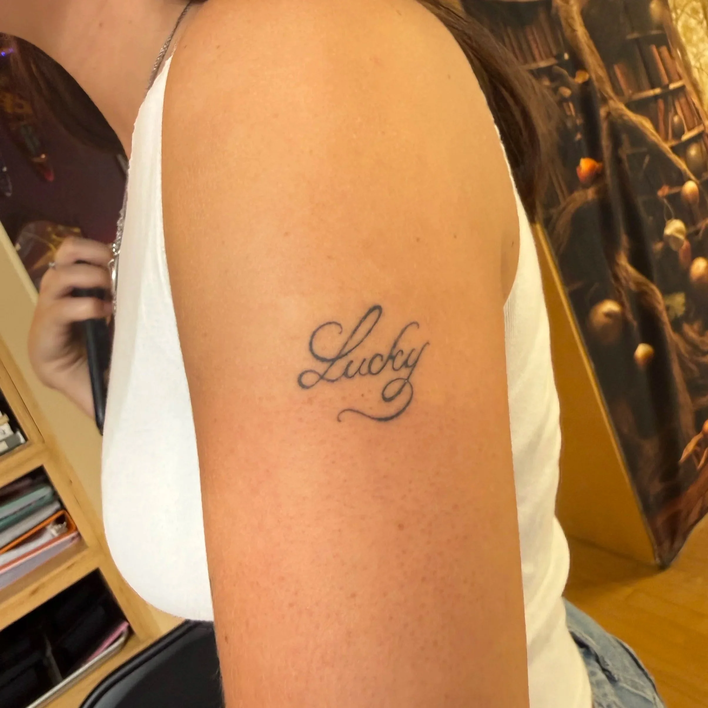 Close-up of a person's back with a tattoo that says 'Lucky'. The person is wearing a white top and a necklace with a small pendant. In the background, there is a bookshelf filled with books and other items.