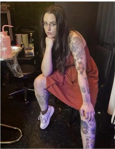 A woman with long dark hair, glasses, and tattoos on her arms and legs sitting on a black chair, leaning forward with her hand on her chin, wearing a sleeveless dress and sneakers, in a room with a black background and a small table with a large pink