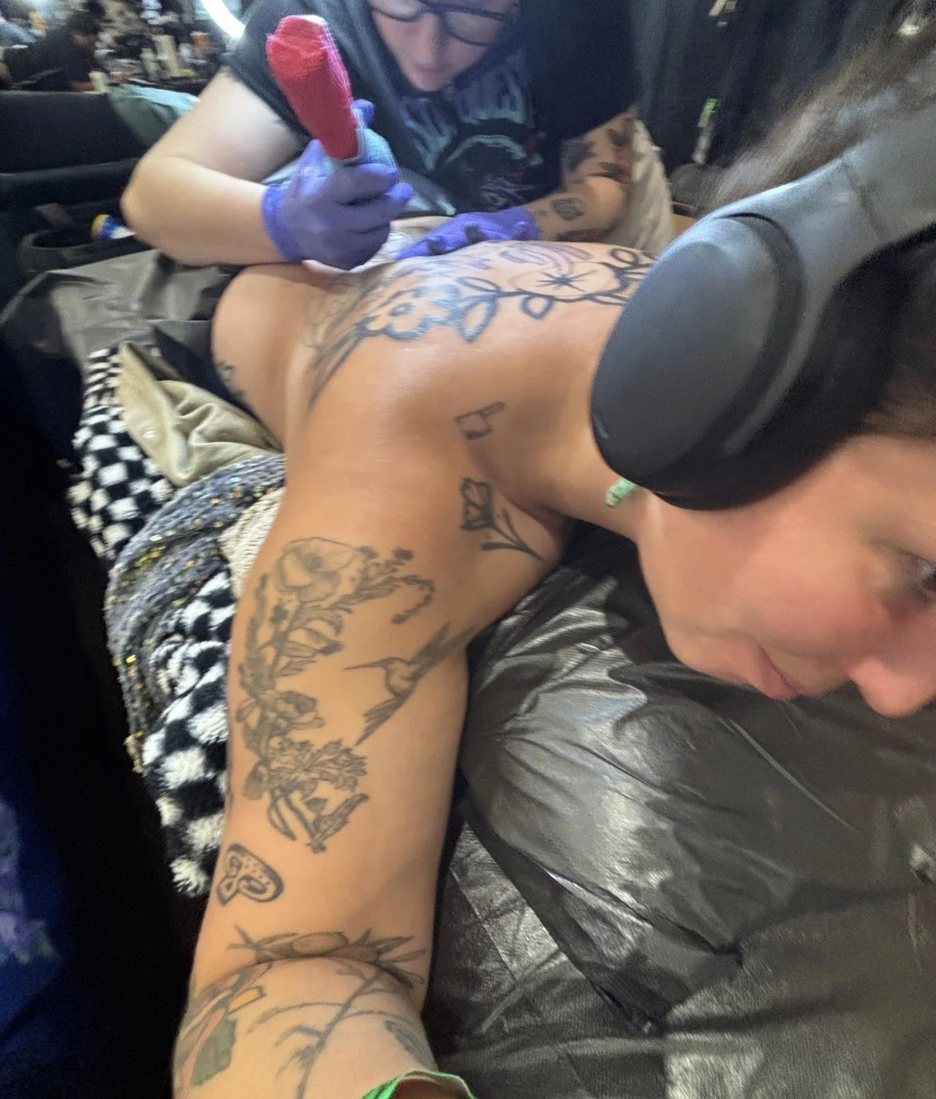 A person with tattoos and wearing headphones is lying face down, getting a tattoo on their back from an artist wearing gloves in a tattoo studio.